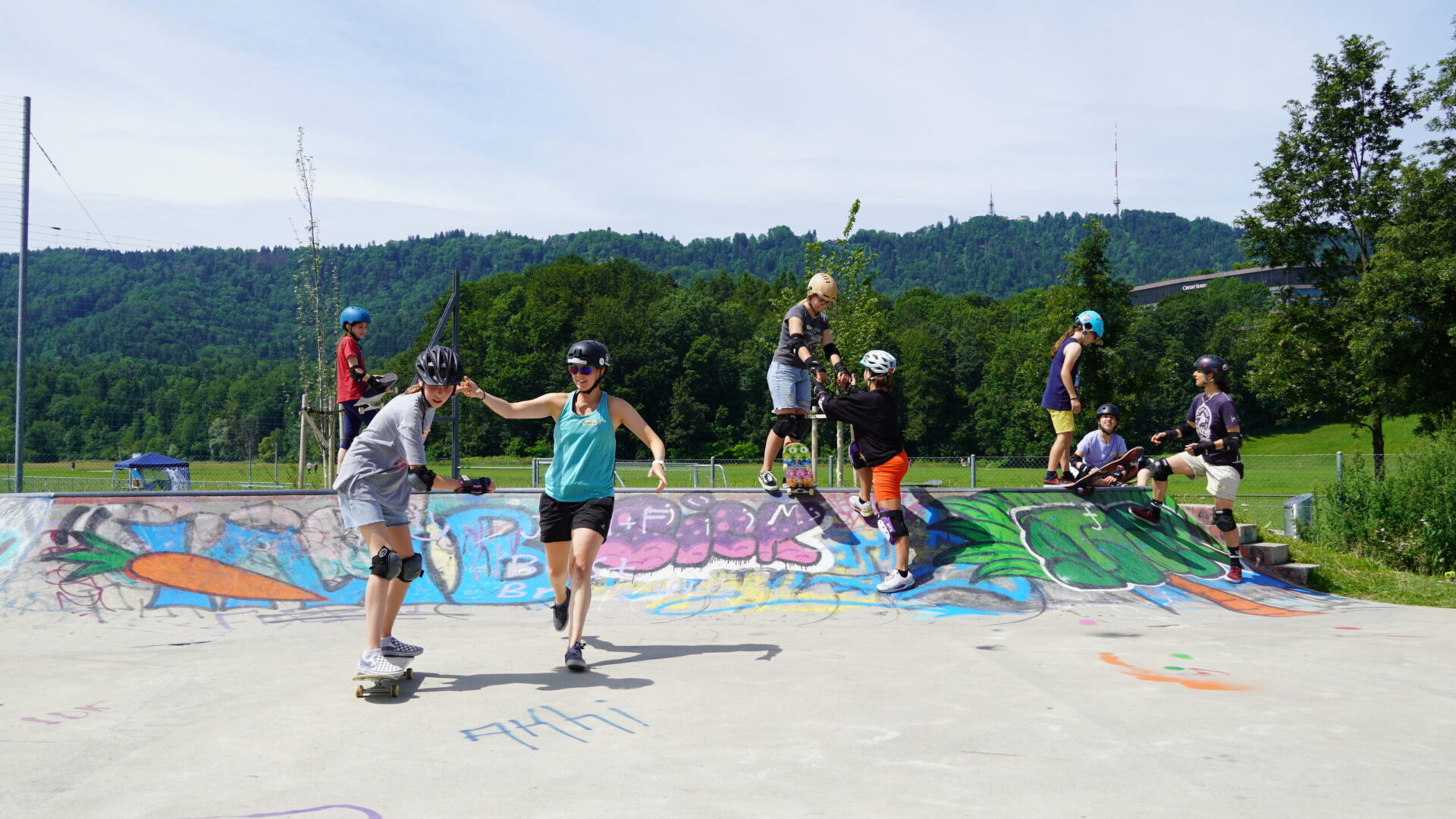 Skaters enjoying a sunny day at a graffiti-covered skate park, practicing moves with the backdrop of a lush green hill.