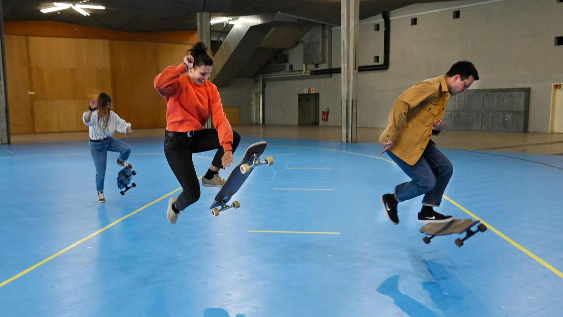 Skaters practicing tricks indoors on a blue floor, captured mid-air during jumps, LGC Switzerland.